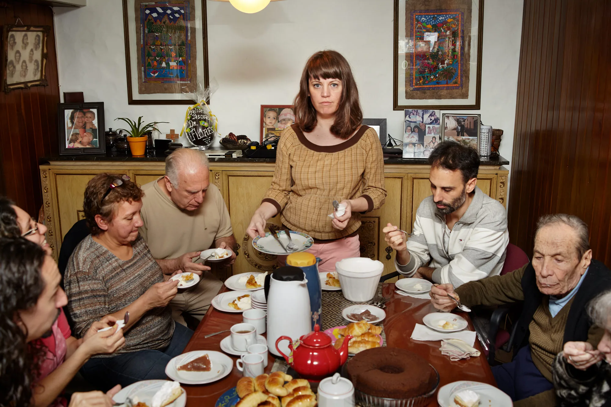 A Woman serves dessert at a crowded Flowers family gathering table, surrounded by relatives enjoying coffee, cake, and pastries in a cozy dining room decorated with framed photos.The Flowers Family documentary series, Argentina