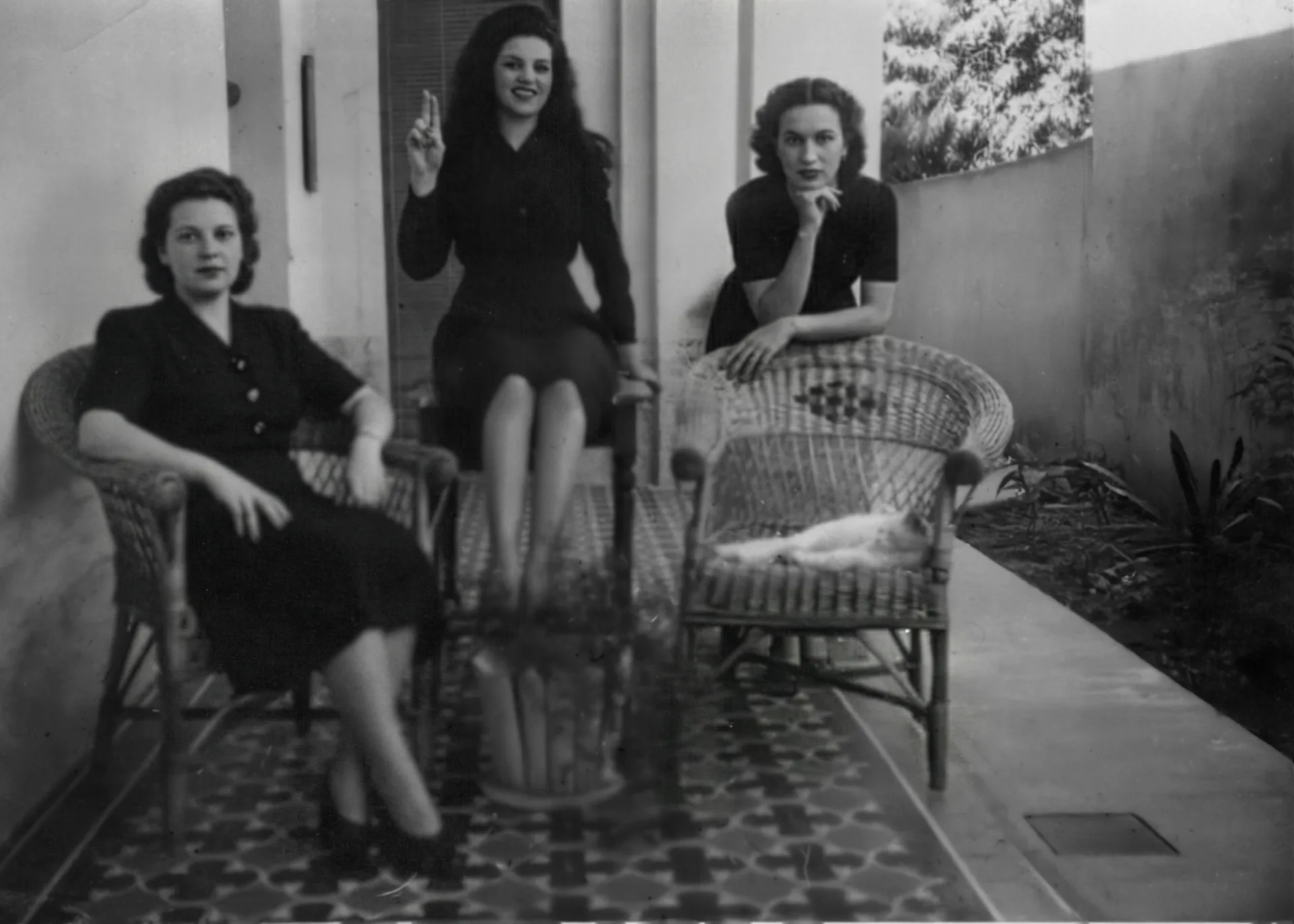 Black-and-white vintage photo of three women posing on a tiled patio with wicker chairs; one sits with crossed legs, one perches on a chair holding up two fingers, and one leans forward with her chin on her hand, while a cat lies on a wicker chair beside them. Disaffiliation series, 2025–2026.
