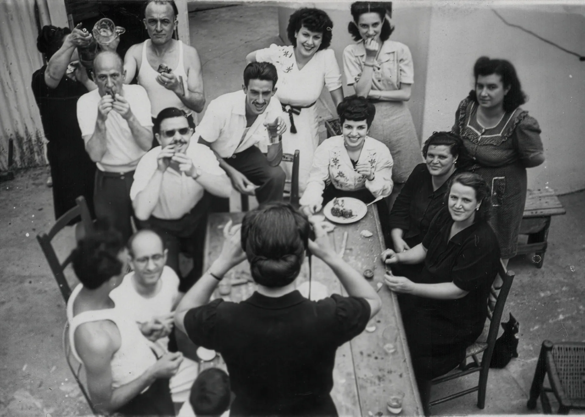 Family Gathering.Black-and-white overhead photo of a lively outdoor gathering around a long table, with a woman seen from behind holding a camera as men and women look up smiling, eating, drinking, and smoking in a courtyard. Disaffiliation series, 2025–2026.