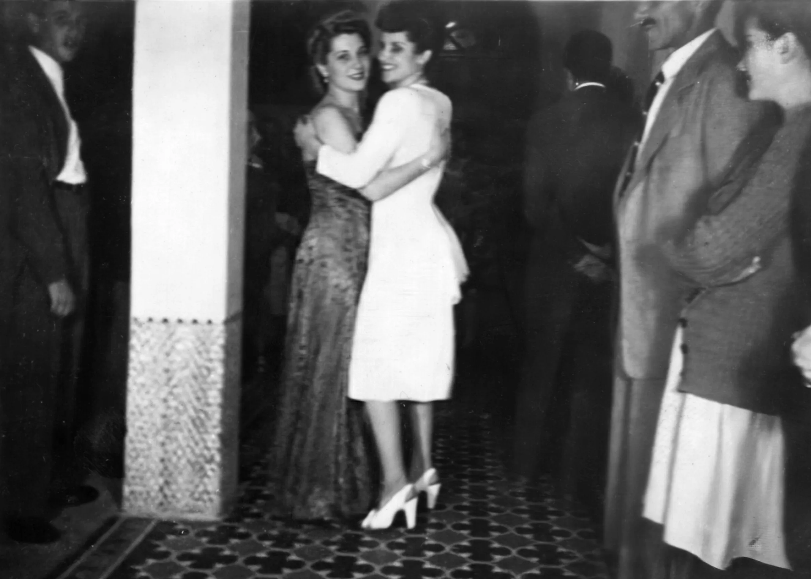 Two women embrace and dance for the camera at an indoor party, framed by other guests along the edges and a patterned tile floor in a black-and-white vintage photograph. Disaffiliation series, 2025–2026.