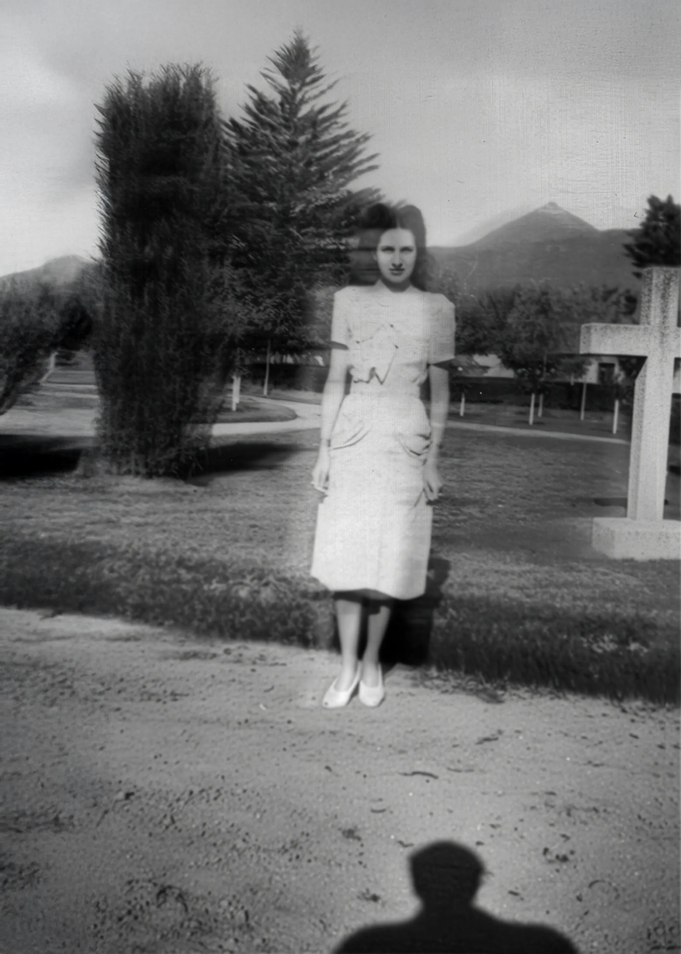 Black-and-white vintage photo of a woman in a light dress standing outdoors on a lawn, her face slightly blurred as if double-exposed, with tall trees, distant mountains, and a large cross to the right; the photographer’s shadow falls in the foreground. Disaffiliation series, 2025–2026.