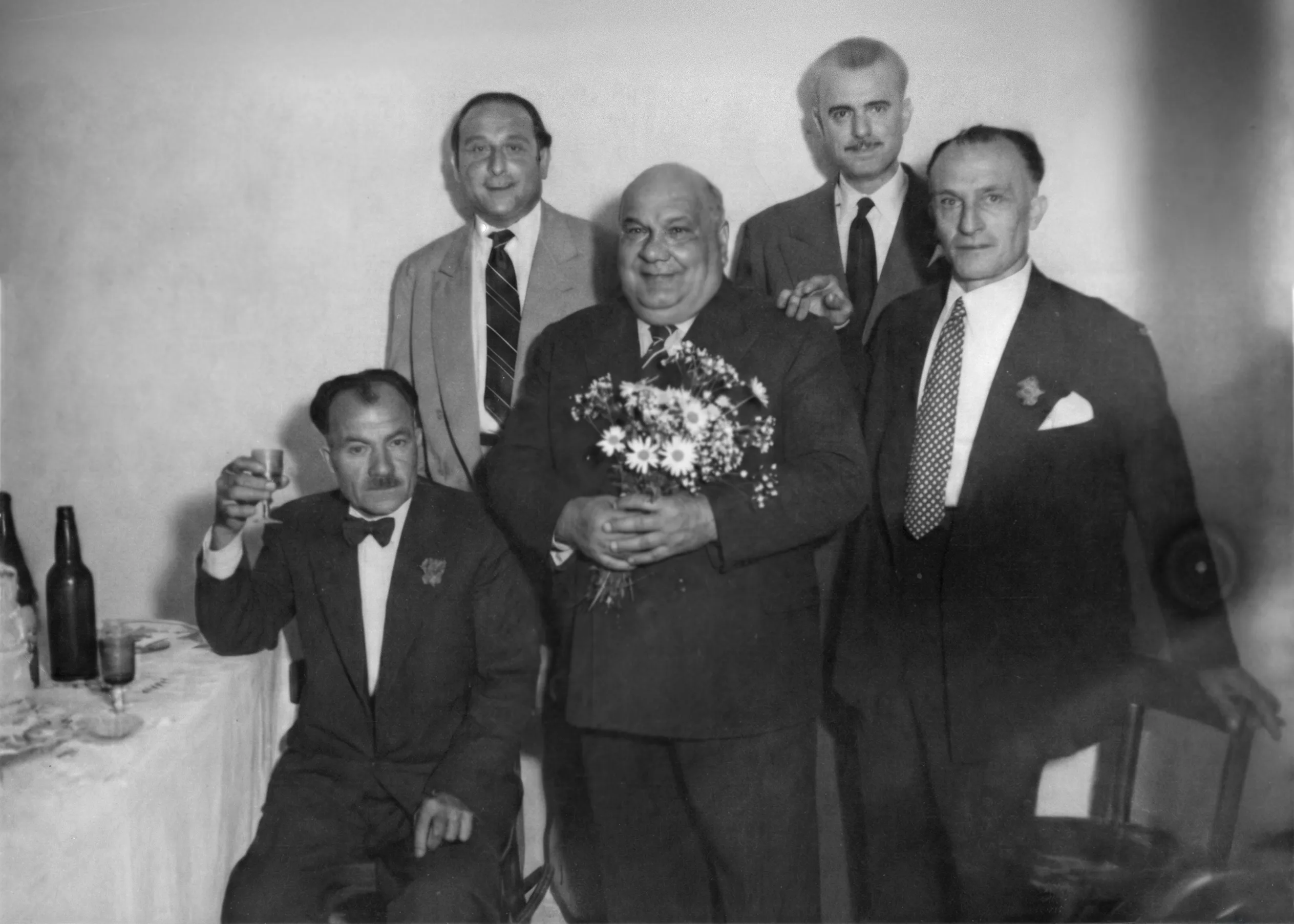 Black-and-white indoor portrait of five men in suits: one seated at a table raising a drink, while a heavyset man stands in the center holding a bouquet of daisies, with three other men standing behind them against a plain wall. Disaffiliation series, 2025–2026.