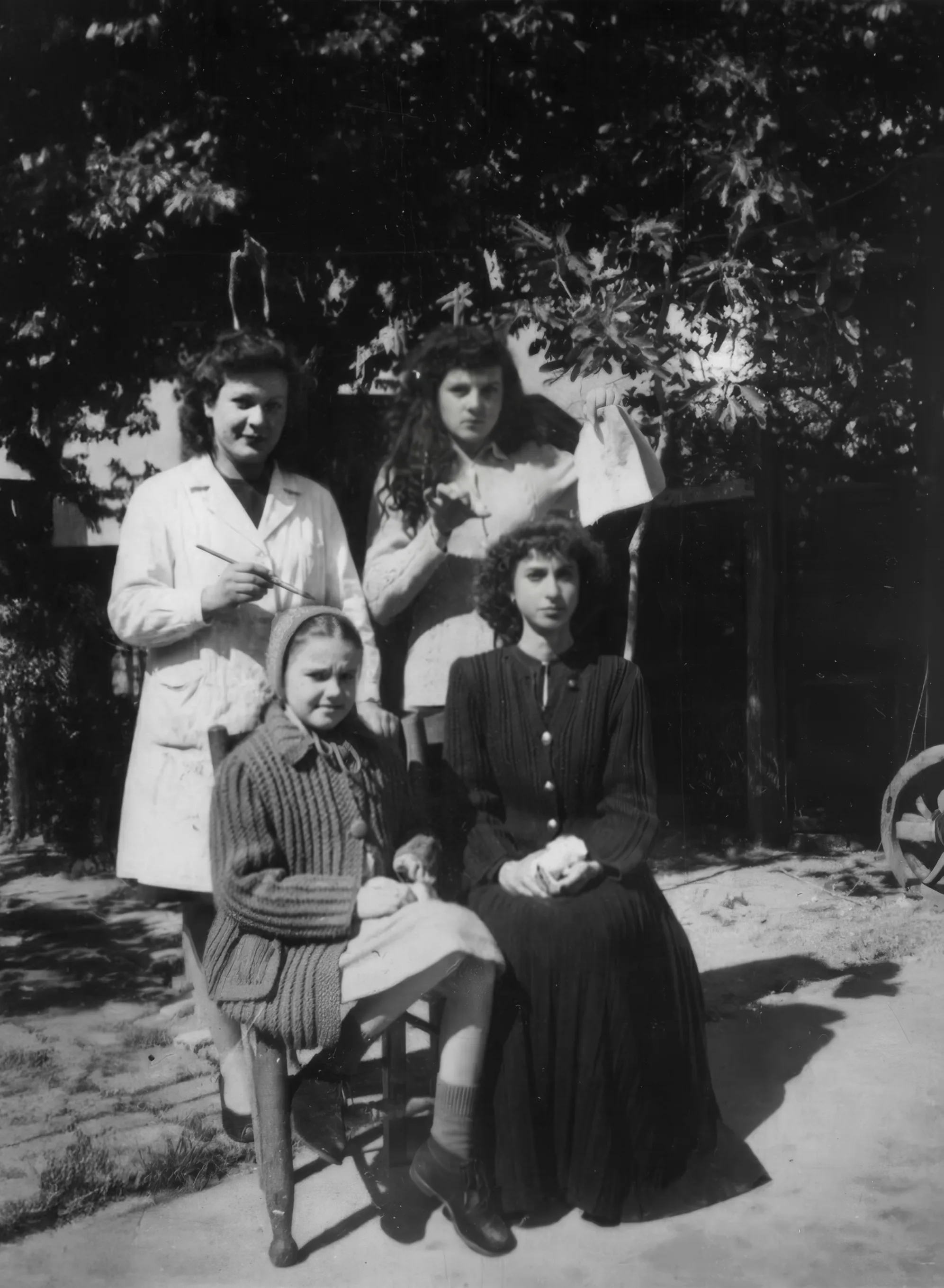 Black-and-white photo of four females posed outdoors under a leafy tree: two seated (a girl in a headscarf and knit cardigan, and a young woman in a dark buttoned dress) with two standing behind, one in a light coat holding a thin brush and the other lifting a cloth, in dappled sunlight near a rustic structure. Disaffiliation series, 2025–2026.