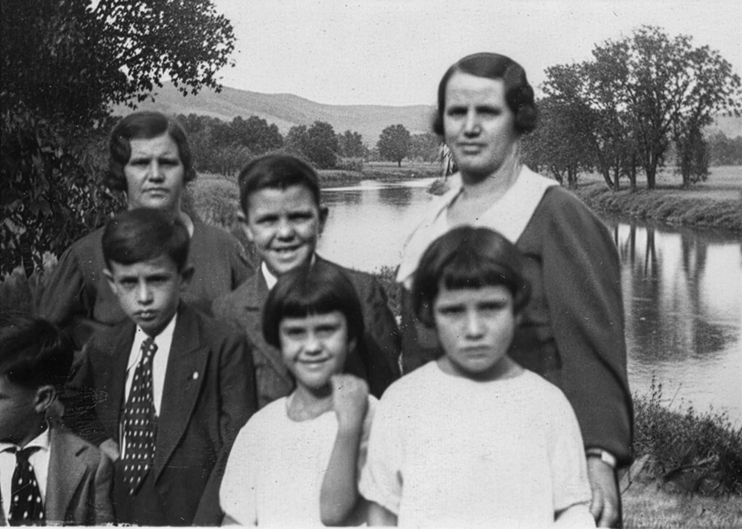 Black-and-white vintage family photo of two women and several children posed by a river, with trees and low hills in the background. Disaffiliation series, 2025–2026.
