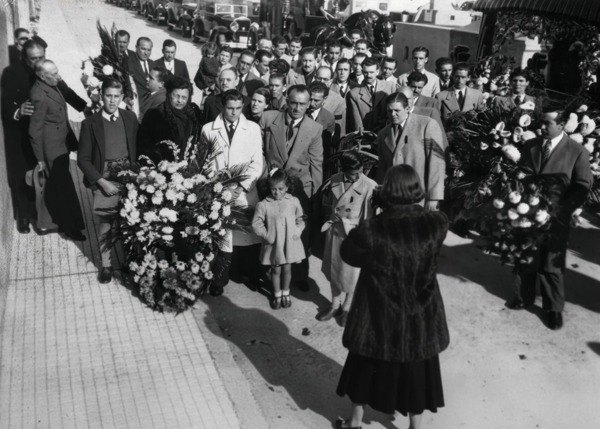 Black-and-white vintage photo of mourners gathered at a cemetery, standing around large floral wreaths and a hearse, while a woman seen from behind in a dark coat raises a camera to photograph the group; two children stand near the front. Disaffiliation series, 2025–2026.