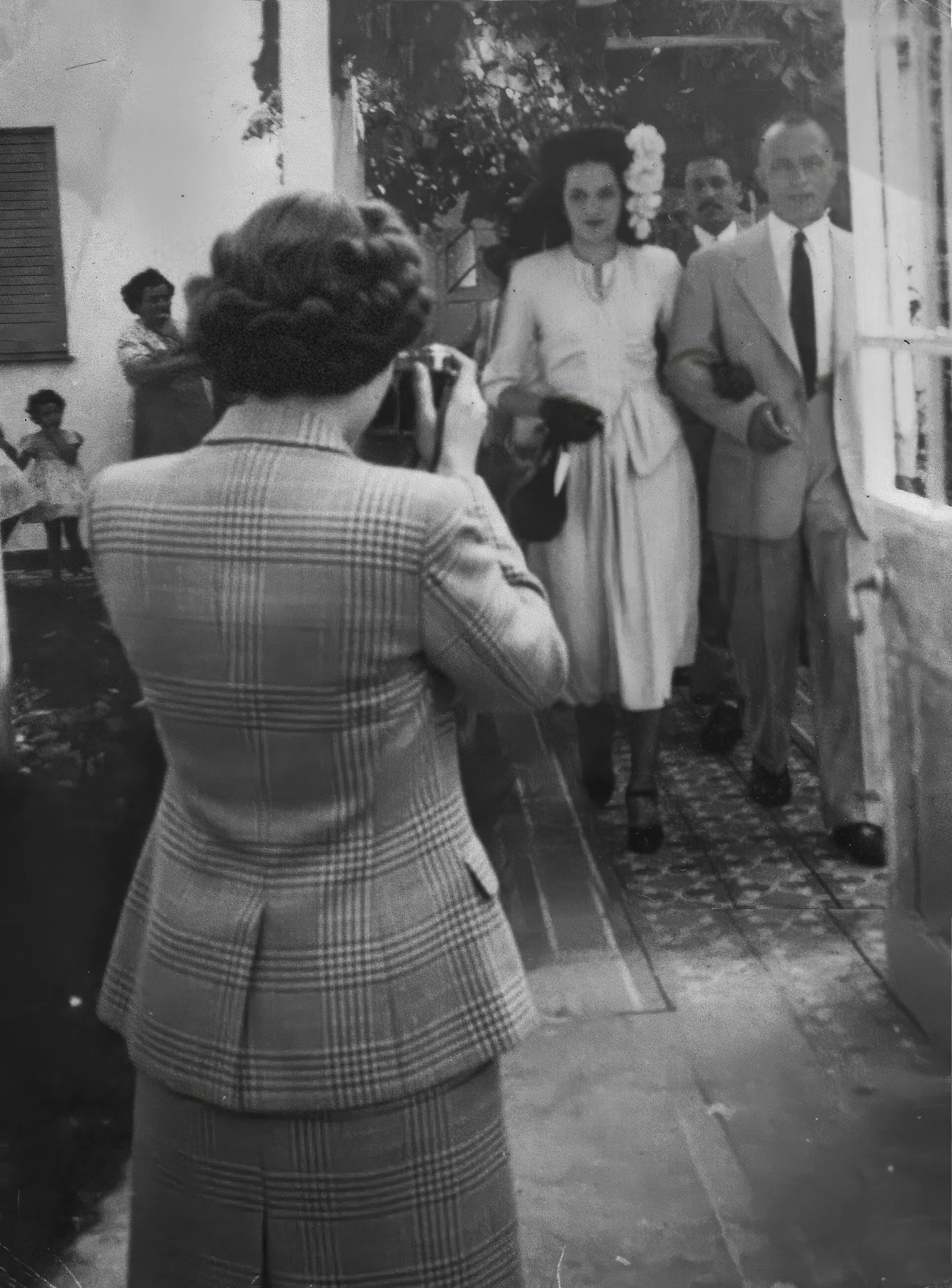 The Wedding Photographer. Black-and-white vintage photo of a woman seen from behind holding a camera, photographing a well-dressed couple walking toward her along a narrow outdoor walkway. Disaffiliation series, 2025–2026.
