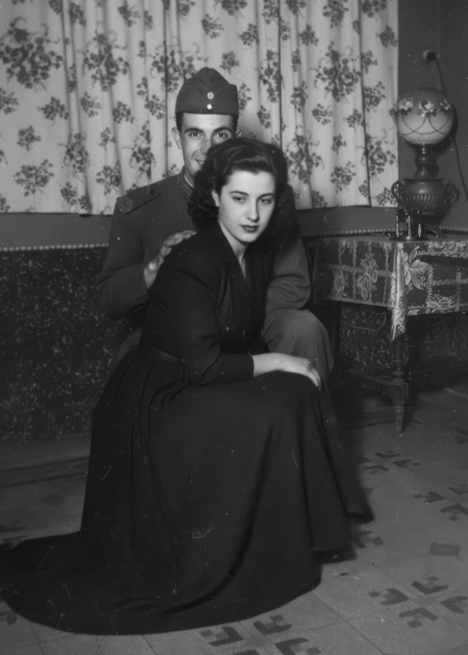 Black-and-white vintage portrait of a woman in a long dark dress seated in front of a uniformed soldier who poses behind her on a sofa covering part of his face, with floral curtains and an oil lamp on a side table.Disaffiliation series, 2025–2026.