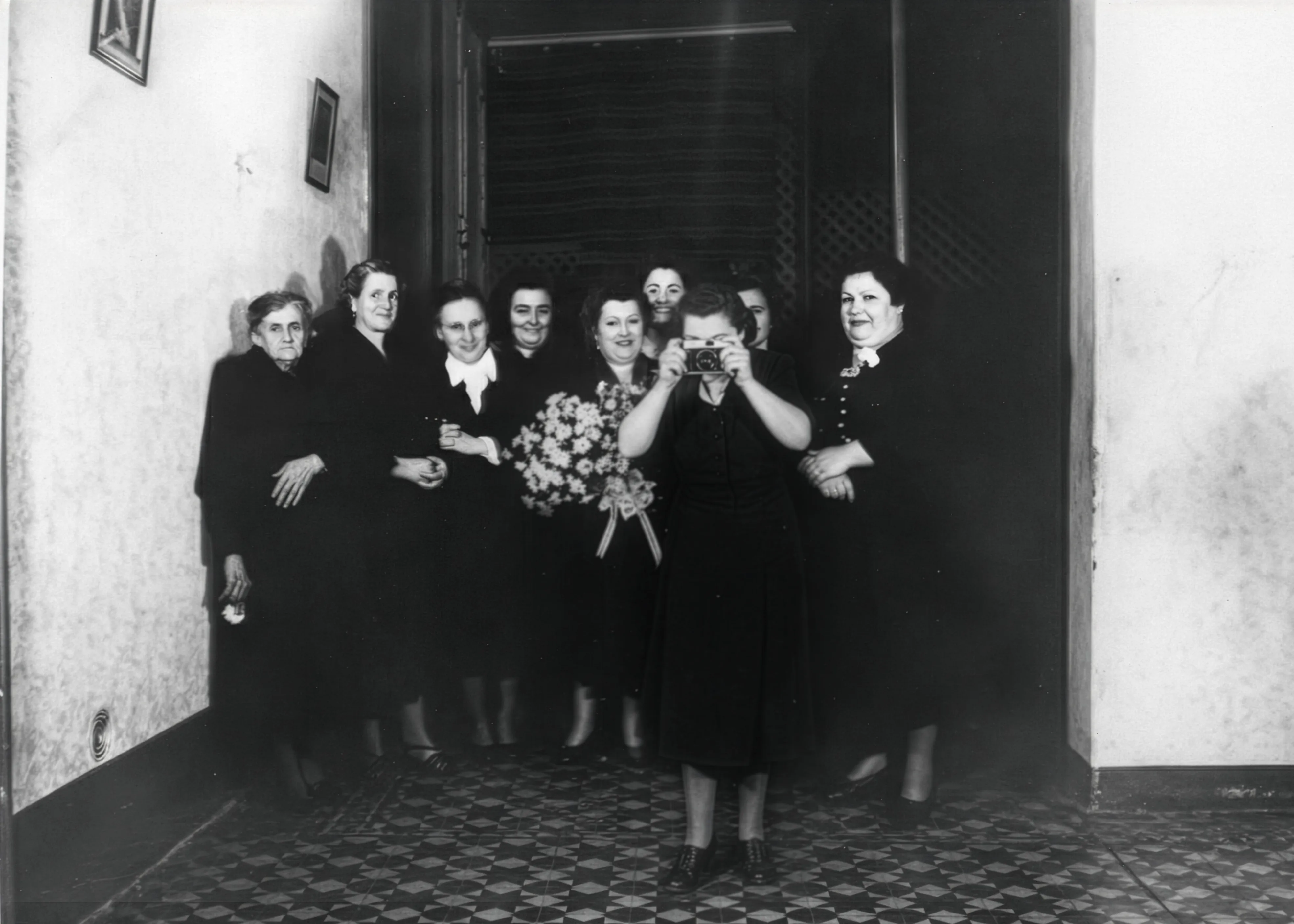 A Female Photographer. Black-and-white vintage photo of a group of women in dark dresses standing in a doorway; one woman in front holds a camera up to her face as if photographing the viewer, while another holds a bouquet.Disaffiliation series, 2025–2026.